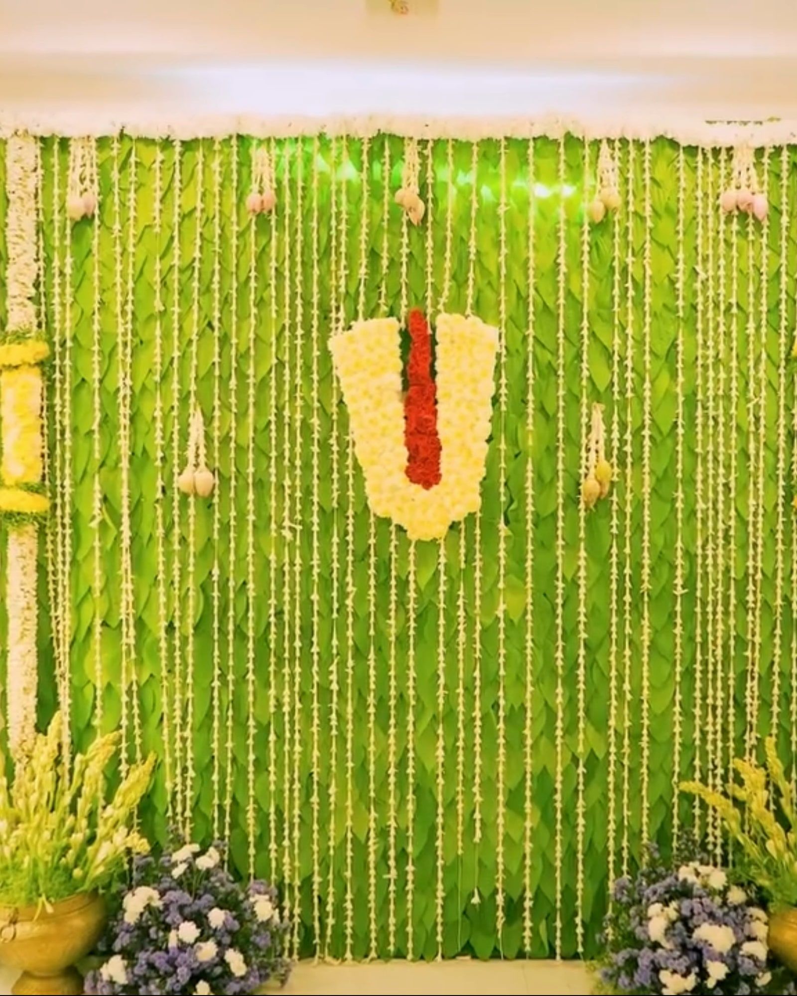 Pooja wall covered in betel leaves and jasmine strings with a flower namam at the centre