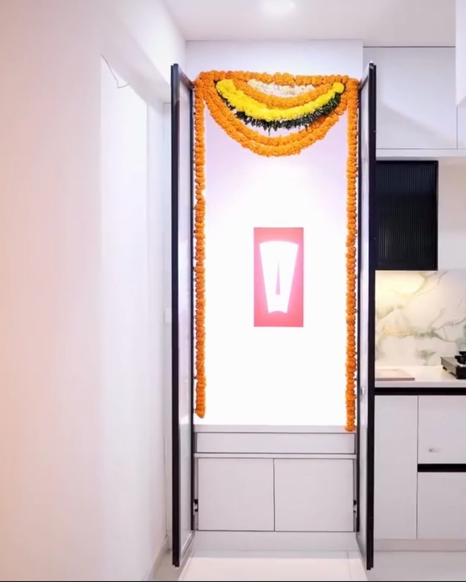 White pooja niche with a marigold toran, framed by a doorway, with the kitchen beyond