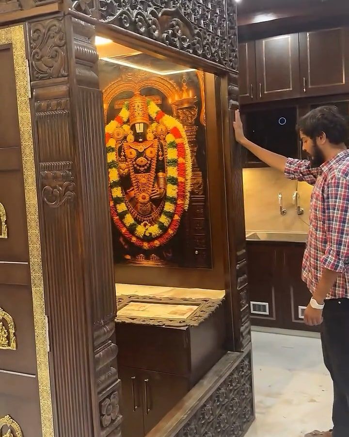 A man rests a hand on the carved pooja temple, a garlanded deity image lit inside