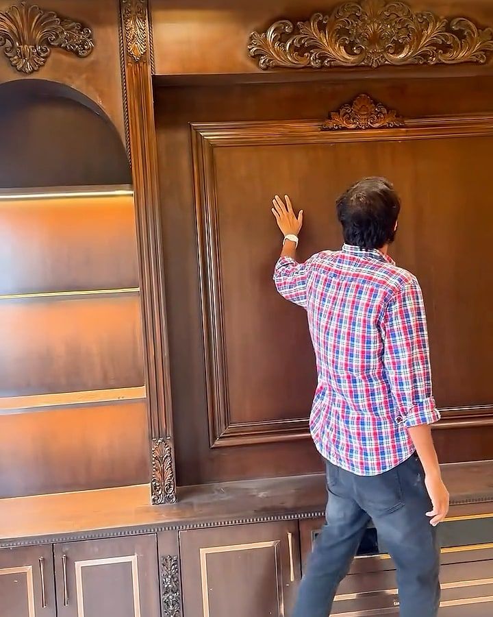 A man in a checked shirt touches the panelled TV wall, carved crests above and lit arched shelves beside it